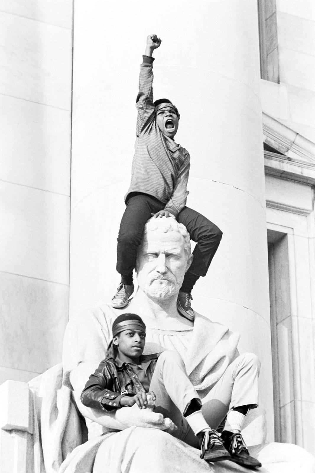Boy gives raised fist salute as he and a friend sit on a statue in front of the courthouse during the Bobby Seale trial.